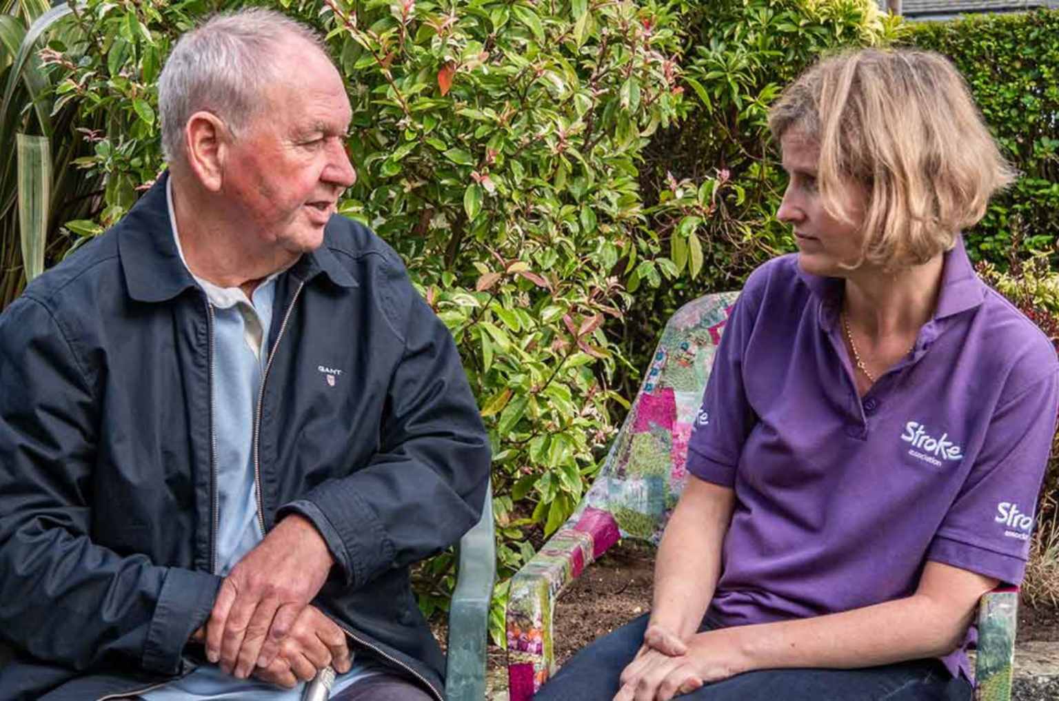 A Stroke Association support worker in a purple polo talking with an elderly man in a garden